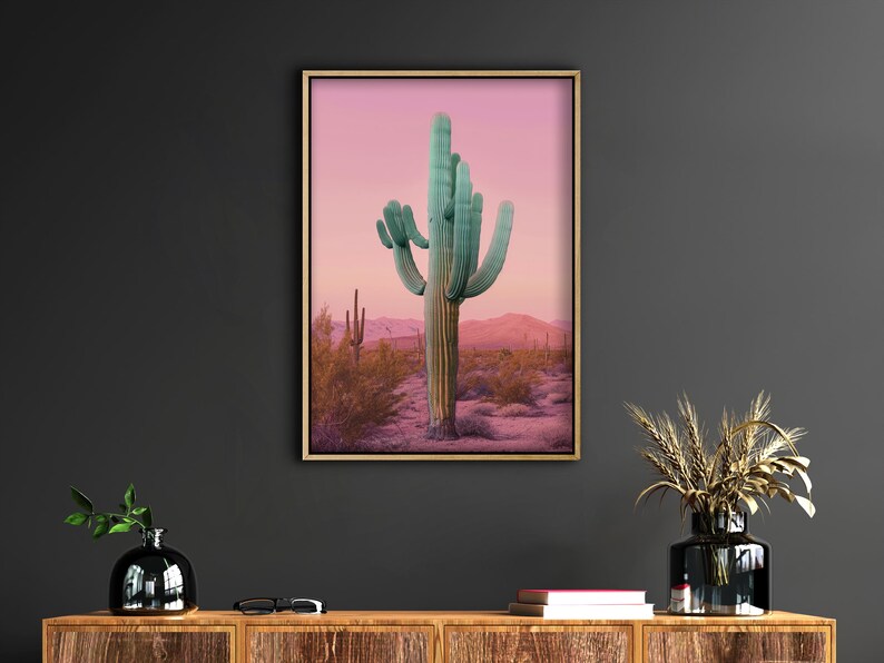 Może przedstawiać: Oprawiony wydruk przedstawiający kaktus saguaro na pustynnym krajobrazie. Kaktus ma żywy turkusowy kolor, osadzony na r&oacute;żowo-fioletowym niebie o zachodzie słońca. Grafika jest wyświetlana na ciemnoszarej ścianie.