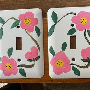 May include: Two white light switch covers decorated with hand-painted pink flowers, yellow centres, and green leaves. The floral design is on a white background. Each cover has a rectangular switch opening and two screw holes.