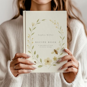 Könnte beinhalten: Ein weißes Rezeptbuch mit einem floralen Kranzdesign und dem Text "Sophia Miller RECIPE BOOK A sprinkle of love in every recipe."