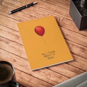 May include: A mustard-coloured notebook featuring a red balloon illustration and the text "ALL I THINK ABOUT IS POOH". A black pen lies on a wooden surface near the notebook. A black mug and a small potted plant are also visible.