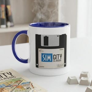 May include: White ceramic mug with a blue handle and rim, featuring a black and white floppy disk design with the text "SIM CITY CLASSIC." Steam rises from the mug. A manual and keyboard keys are also visible.