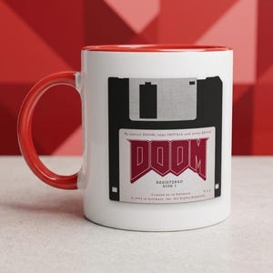 May include: A white ceramic mug with a bright red handle and interior. The front features a graphic of a black floppy disk with a white label that reads DOOM, Registered Disk 1, Created by id Software, V 1.2, and installation instructions.