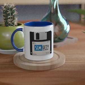 May include: White ceramic mug with a blue interior and handle. The mug features a black and white graphic of a floppy disk with the text "SIM CITY CLASSIC". The mug sits on a wooden coaster.
