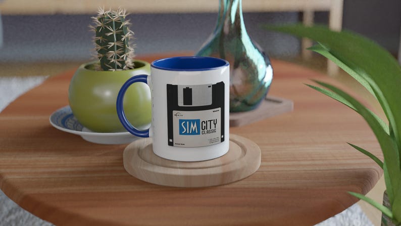 May include: White ceramic mug with a blue interior and handle. The mug features a black and white graphic of a floppy disk with the text "SIM CITY CLASSIC". The mug sits on a wooden coaster.
