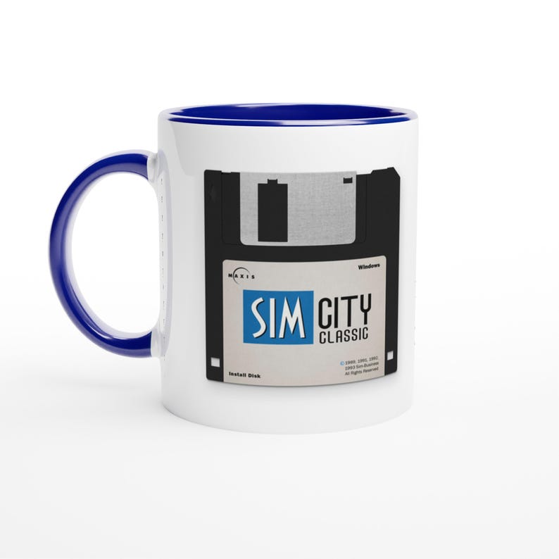 May include: White ceramic mug with a blue interior and handle. The mug features a black and gray design of a floppy disk with the text "SIM CITY CLASSIC" in blue and white.