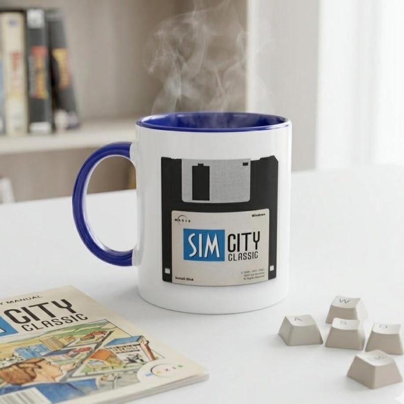 May include: White ceramic mug with a blue handle and rim, featuring a black and white floppy disk design with the text "SIM CITY CLASSIC." Steam rises from the mug. A manual and keyboard keys are also visible.
