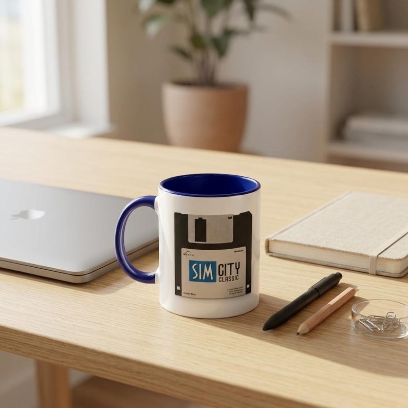 May include: White ceramic mug with a blue interior and handle. The mug features a black and white design of a floppy disk with the text "SIM CITY CLASSIC" in blue and white. The mug is on a wooden surface.
