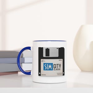 May include: White ceramic mug with a blue handle and rim, featuring a retro design of a floppy disk with the text "SIM CITY CLASSIC". The mug is placed next to a stack of books and decorative vases.