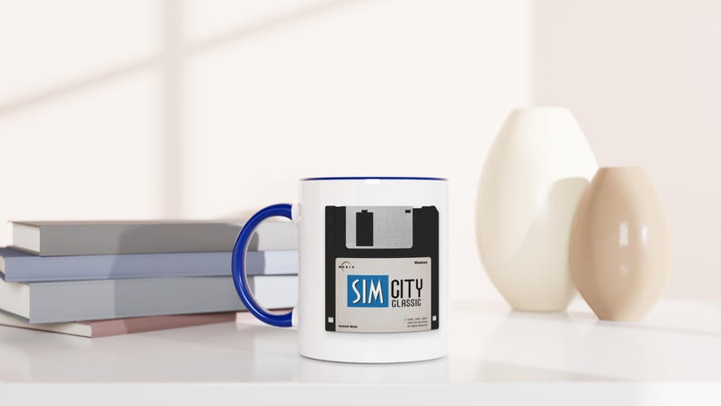 May include: White ceramic mug with a blue handle and rim, featuring a retro design of a floppy disk with the text "SIM CITY CLASSIC". The mug is placed next to a stack of books and decorative vases.