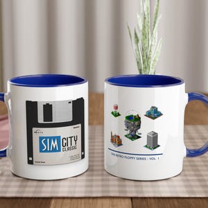 May include: Two white ceramic mugs with blue interiors and handles. One mug features a floppy disk graphic with "SIM CITY CLASSIC" text. The other mug displays retro game graphics and the text "THE RETRO FLOPPY SERIES - VOL. 1."