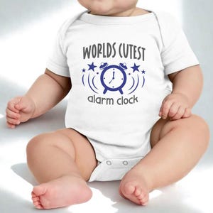 May include: White baby onesie with the text "Worlds Cutest alarm clock" in gray and blue. The design features an alarm clock graphic with stars. The onesie is short-sleeved and has snap closures.