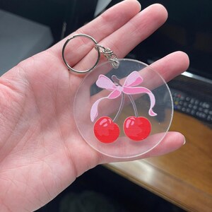 May include: Clear acrylic keychain featuring a pair of red cherries with a pink bow. The keychain has a silver metal ring for attaching keys. The design is a cute, playful accessory.