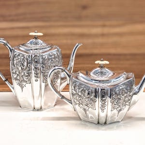 May include: Two silver teapots with a hexagonal shape and intricate floral engravings. Each teapot features a curved handle, a slender spout, and a lid topped with a small, cream-colored circular finial. The metal surfaces are highly reflective.