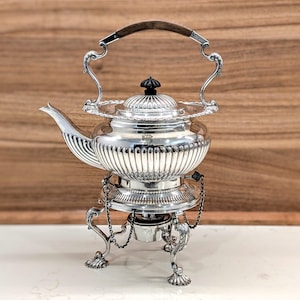 May include: A polished silver tea kettle with a fluted body, a dark wooden handle, and a matching lid with a black finial. The kettle rests on a decorative silver stand featuring three ornate legs, claw feet, and a central burner attachment.