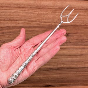 May include: A silver-toned serving fork featuring a long, slender handle with an intricate, embossed floral pattern and a three-pronged head. The metal surface is polished and reflective, showcasing detailed scrollwork along the entire handle.