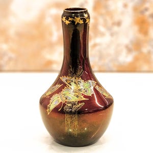 May include: A decorative vase with a dark burgundy and gold finish. The vase features a flared base and a slender neck, adorned with gold star accents and butterfly designs. The butterfly wings are detailed with green and gold accents.