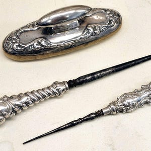 May include: An antique silver shoe brush and two silver tools with ornate handles. The brush is oval with a silver top and a tan brush base. One tool has a hook, and the other has a pointed end. The tools have dark, aged metal.