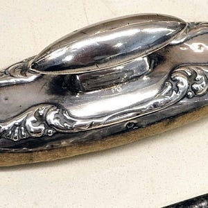May include: An oval-shaped silver-toned desk blotter with a central handle. The top surface features an intricate embossed floral and scrollwork pattern. The base is made of a tan-coloured felt material, providing a flat surface for blotting ink.