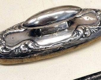 May include: An oval-shaped silver-toned desk blotter with a central handle. The top surface features an intricate embossed floral and scrollwork pattern. The base is made of a tan-colored felt material, providing a flat surface for blotting ink.