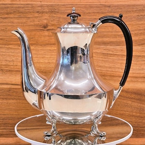 May include: A polished silver tea or coffee pot with a black handle and a decorative finial on the lid. The pot has a curved spout and sits on a mirrored surface. The pot is a classic design.