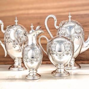 May include: A four-piece silver tea set comprising a large teapot, a smaller teapot, a creamer, and a sugar bowl. Each piece is crafted from polished silver with intricate engraved floral and scrollwork patterns, pedestal bases, and curved handles.