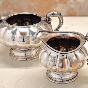May include: A silver sugar bowl and creamer set. The items have a fluted design and decorative handles. The creamer has a spout. The set is likely antique or vintage, with some wear visible on the surface.