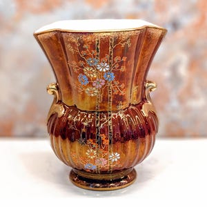 May include: A fluted ceramic vase with a lustrous copper-brown iridescent glaze, featuring two gold-toned scroll handles and a delicate hand-painted floral pattern in blue, orange, and white. The interior of the vase is finished in white.