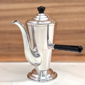 May include: A silver-toned metal coffee pot with a tapered body, a curved spout, and a circular base. The pot features a stepped lid topped with a small black finial and a horizontal black handle extending from the side, providing a contrast to the metallic finish.
