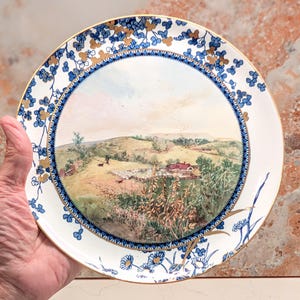 May include: A round ceramic plate featuring a central landscape illustration of a rural hillside with a house, trees, and a person walking a dog. The border is decorated with blue floral patterns and gold accents on a white background.