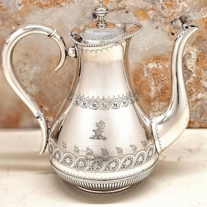 May include: A polished silver coffee pot with a curved handle and a long, elegant spout. The pot has intricate decorative patterns around the body and lid. The pot is made of a shiny metal, and is a classic design.