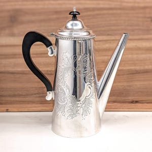 May include: A silver-colored coffee pot featuring an intricate engraved floral pattern on the body, a tall tapered spout, and a curved black handle. The lid is topped with a small black finial, and the base of the lid has a beaded metal trim.