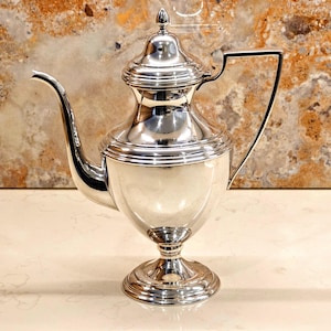 May include: A polished silver coffee pot with a curved spout and a handle. The pot has a decorative lid with a small finial. The base is a pedestal design. The pot is likely made of stainless steel or silver.