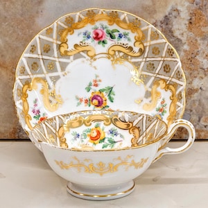 May include: A white teacup and saucer set with gold trim and floral designs. The saucer has a grey and gold lattice pattern. The cup and saucer feature colourful flowers and gold accents, creating an elegant and vintage aesthetic.