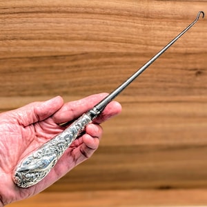May include: A silver ear spoon with a decorative handle and a hooked end. The handle has detailed floral and scrollwork designs. The spoon is held against a wooden background.