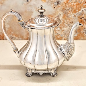 May include: A silver teapot with a curved handle and a decorative spout. The teapot has a ribbed body and a lid with a small finial. The teapot sits on ornate feet. The background is a mottled brown and beige.