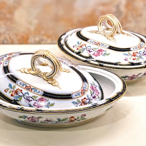 May include: Two white porcelain serving dishes with lids. Each dish features a floral pattern with pink, orange, and blue flowers, a black border, and gold accents. The lids have gold handles.