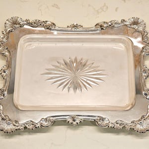 May include: A silver serving tray with a decorative, ornate border and a clear glass insert with a starburst pattern.