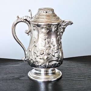 May include: Ornate silver-plated coffee pot with a lid and handle. The pot features detailed floral and scrollwork designs. The spout is adorned with a sculpted face. The pot sits on a round base and is displayed on a dark wooden surface.