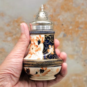 May include: A ceramic shaker with a silver metal top. The shaker has a floral design in orange and blue against a cream background. The top has a pierced design. The shaker is held in a hand.