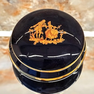May include: A navy blue trinket box with a gold trim and a gold illustration of a couple in historical attire. The box has a glossy finish and is reflected in the surface below.