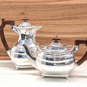 May include: A polished silver coffee pot and a matching silver teapot, both featuring dark brown wooden handles and finials on their lids. Each piece has a rounded, multi-faceted body, a curved spout, and rests on four small, rounded silver feet.