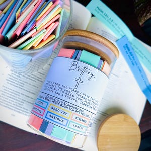 May include: A glass jar filled with colorful paper strips containing Bible verses. The jar has a wooden lid and a label that says "Brittany." The label also includes a cross and the text "FOR I KNOW THE PLANS I HAVE FOR YOU DECLARES THE LORD, PLANS TO PROSPER YOU AND NOT TO HARM YOU. PLANS TO GIVE YOU HOPE AND A FUTURE. JEREMIAH 29:11." The jar is sitting on a Bible.