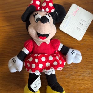 Puede incluir: Un peluche de Minnie Mouse. La mu&ntilde;eca tiene un vestido rojo con lunares blancos, un lazo a juego y zapatos amarillos. El juguete tiene orejas, brazos y piernas negros y guantes blancos. Una etiqueta de Disney est&aacute; adjunta.