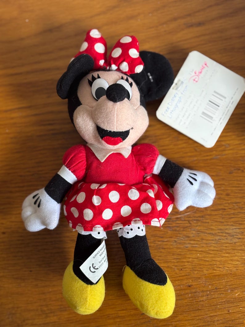Puede incluir: Un peluche de Minnie Mouse. La mu&ntilde;eca tiene un vestido rojo con lunares blancos, un lazo a juego y zapatos amarillos. El juguete tiene orejas, brazos y piernas negros y guantes blancos. Una etiqueta de Disney est&aacute; adjunta.