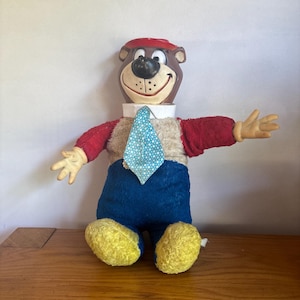 Puede incluir: Un muñeco de peluche del personaje Yogi Bear con cabeza de plástico moldeado marrón, sombrero rojo, cuello blanco y corbata azul claro con lunares. El cuerpo es de tela texturizada con mangas rojas, pecho beige, pantalones azules y pies amarillos.
