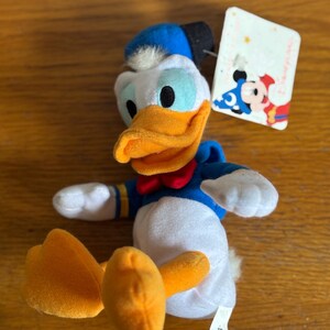 Puede incluir: Un peluche de Pato Donald con camisa de marinero azul, pajarita roja y pico y pies naranjas. El juguete tiene un sombrero azul y un cuerpo blanco. Una etiqueta de Disney est&aacute; adjunta.