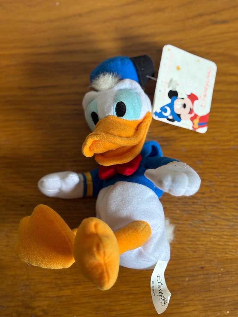 Puede incluir: Un peluche de Pato Donald con camisa de marinero azul, pajarita roja y pico y pies naranjas. El juguete tiene un sombrero azul y un cuerpo blanco. Una etiqueta de Disney est&aacute; adjunta.