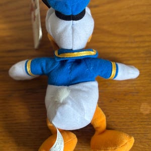 Puede incluir: Un peluche de Donald Duck, visto desde atr&aacute;s, con gorro y chaqueta de marinero azules, cuerpo blanco y pies naranjas. El juguete tiene una etiqueta adjunta. El fondo es una superficie de madera.