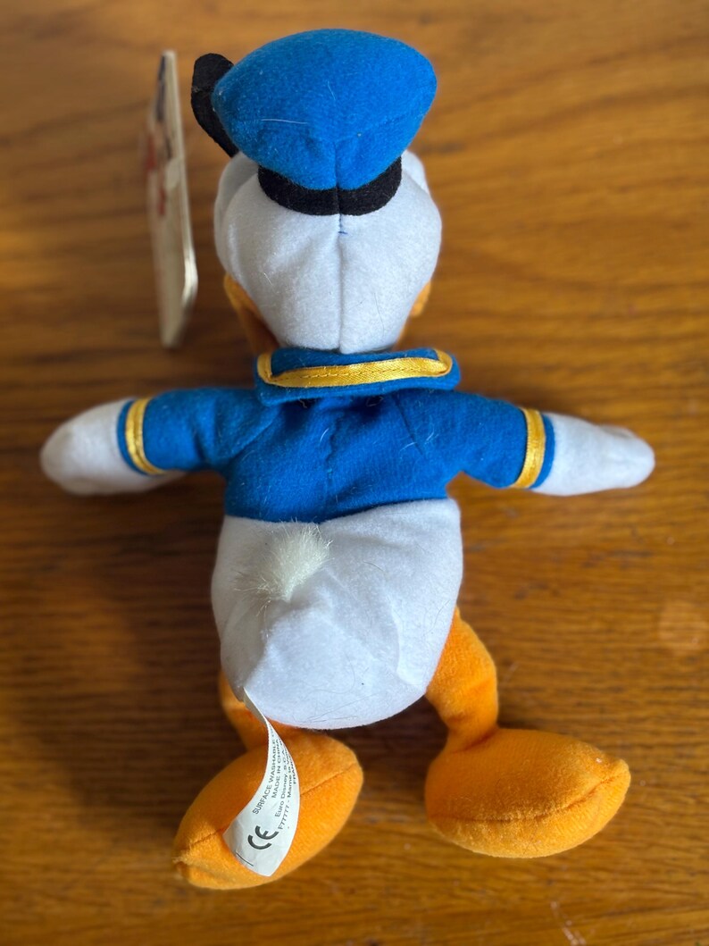 Puede incluir: Un peluche de Donald Duck, visto desde atr&aacute;s, con gorro y chaqueta de marinero azules, cuerpo blanco y pies naranjas. El juguete tiene una etiqueta adjunta. El fondo es una superficie de madera.
