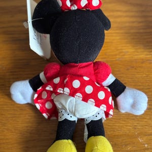 Puede incluir: Mu&ntilde;eca de peluche de Minnie Mouse, vista desde atr&aacute;s, con un vestido y un lazo rojos, ambos adornados con lunares blancos. La mu&ntilde;eca tiene orejas, brazos y piernas negros, con guantes blancos y zapatos amarillos. Una etiqueta est&aacute; adjunta.